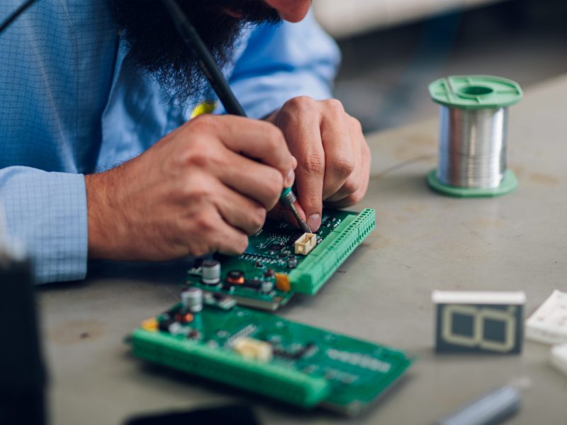 Technician in an antistatic coat repairs circuit board of a device with iron soldering and tin wire. Electronics engineer working in his workshop. Focus on a man hands repairing the device.